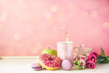Beautiful breakfast. Pink icing donut, macarons, sugar, pink tea cup, green apple, bouquet with yellow flowers, tea kettle on a pink background. Copy space.