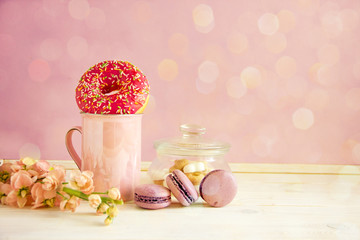 Beautiful breakfast. Pink mug with a handle, sugar bowl, donates with pink icing, macarons cakes, fresh flowers, green apple and a transparent teapot of tea on a pink background. Copy space