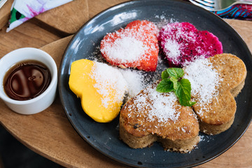 Heart shaped pancakes and tropical fruits on a black plate on wooden table, saint valentine