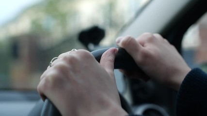 Two female hands on a car steering wheel driving through a suburb in the middle of the day. A joyful drive through the town with greenery and nice houses in the background