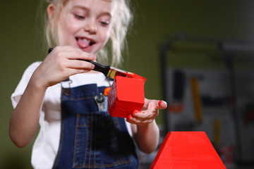 The little girl in the carpentry workshop paints with red paint wooden rectangles, triangle panes,...