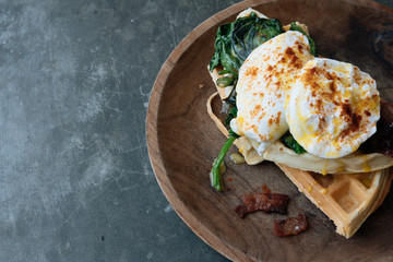 Breakfast with Poached Egg on waffle with spinach and bacon on wooden texture plate on grey stone table. Top view. Space for text