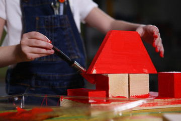 Hands of a little girl who paints with a red paint a wooden house. Photos in the carpentry workshop. The face is out of focus. Macro photo. Concept of development and learning.