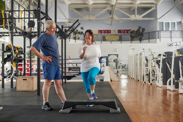 Fototapeta na wymiar Positive senior couple exercising at gym. Happy elderly woman training her legs at fitness club. Keep your body in shape.