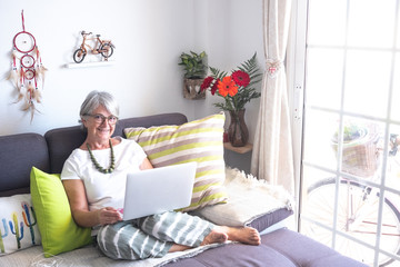 Senior woman white hair sitting on sofa and using laptop. One adult people, caucasian. Bright light from window. Happy and modern. White background