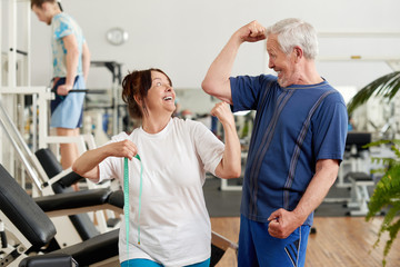 Fototapeta na wymiar Cheerful senior couple showing their biceps at the gym. Joyful senior woman with measuring tape at fitness club. People, sport, leisure and healthy lifestyle concept.