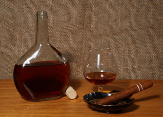 Bottle and wineglass with cognac, white ash tray and cigar on the table with canvas background