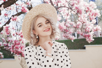 Outdoor fashion portrait of young beautiful happy smiling lady wearing  trendy straw hat, pearl earrings, white wrist watch, blouse, posing in street with blooming magnolia tree. Copy, empty space