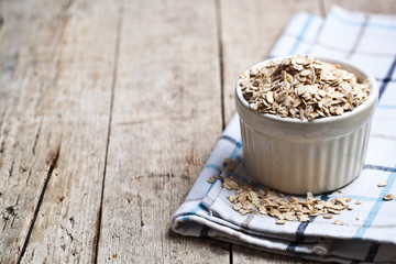 Oat flakes in ceramic bowl on linen napkin, golden wheat ears on rustic wooden background.