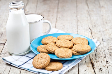 Fresh baked oat cookies on blue ceramic plate on linen napkin, bottle of milk and ceramic cup on rustic wooden table background.