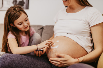Young pregnant woman enjoying wihile her cute daughter applying moisturizer in smiley face shape on her belly.
