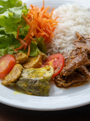 White plate with stewed fish, pork, rice, lettuce, tomato and carrot. Healthy meal served in terrace. Close up. Daylight. 