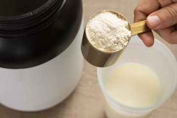 Whey Protein. Shaker with milk. Perspective, holding measuring scoop full of vanilla flavour powder on wooden background.