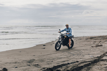 Fototapeta na wymiar Man in cap riding motorcycle on beach. Moto cross dirtbiker on beach sunset on Bali. Young hipster male enjoying freedom and active lifestyle, having fun on a bikers tour.