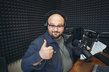 Funny happy radio presenter or host in radio station studio looks at camera and shows thumbs up...