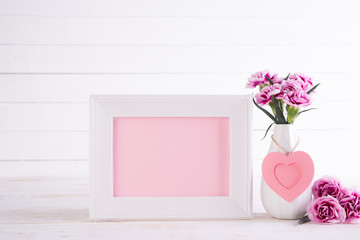 White picture frame with lovely pink carnation flower in vase on white wooden table.