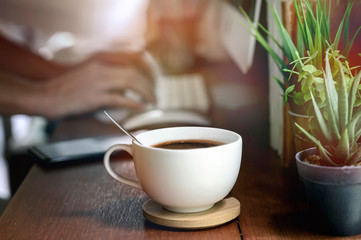 Closeup image of white cup of coffee on wooden teable with blur image of hand typing computer keyboard.