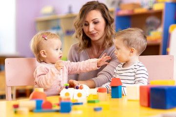 Babies with mentor in kindergarten. Kids toddlers in nursery school. Little girl and boy preschoolers playing with teacher. - 267500767
