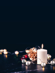 Christmas white candle with golden pine cone,mistletoe and light string glowing on black marble...