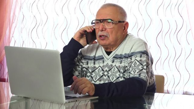 Senior Man With Wrinkly Face Puts Down Information Into Silver Laptop Takes Black Phone And Talks Closeup