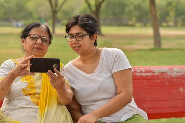 Young Indian girl with her old Indian mother looking at the mobile phone and busy talking selfie on a red bench in a park in New Delhi, India. Concept Mother's day