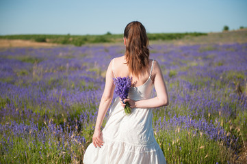 Fototapeta na wymiar young beautiful elegant female in white dress holding bouquet of lavender flower behind her back standing in bloom field outdoors leisure lifestyle