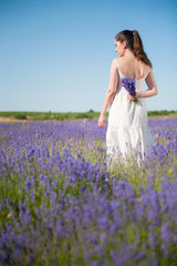 Fototapeta na wymiar young alone elegant caucasian woman in white dress holding bouquet of lavender flower behind her back standing in flowering summer field outdoor lifestyle