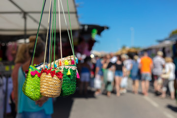Many colorful wicker basket bags hang on a stall at a weekly market in Spain. In the background you can see people looking at the goods.