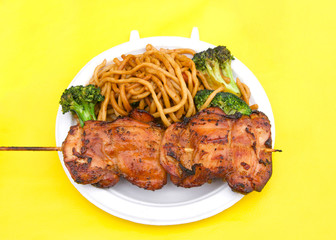 White paper plate with teriyaki chicken on skewer with chow mein noodles on yellow table cloth. Popular street fair food.