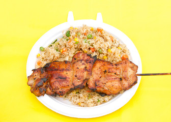 White paper plate with teriyaki chicken on skewer with fried rice on yellow table cloth. Popular street fair food.