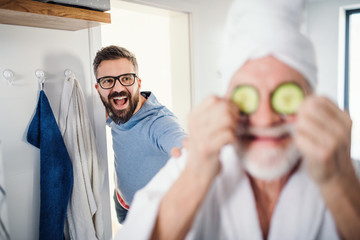 An adult hipster son and senior father in bathroom indoors at home, having fun.