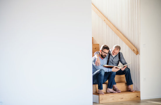 An Adult Son And Senior Father Sitting On Stairs Indoors At Home, Looking At Photographs.