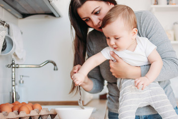 Young mother brunette woman with baby boy in arms cooking food in white modern kitchen at home