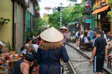 Fototapeta na wymiar Foreign tourist with Vietnamese conical hat walking on railroad in Hanoi. People drink coffee or walking on railways waiting for train to arrive on railway road on background in Hanoi, Vietnam.