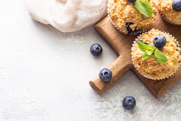 Blueberry muffins with fresh berries and mint leaves on a wooden board. Light grey stone background. Copy space.