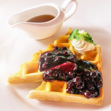 High key image of a beautiful Belgian waffle served with blueberry compote and whipped cream, garnished with a mint leaf. A serving dish of maple syrup sits on a white plate background.