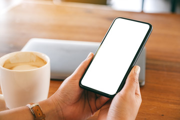 Mockup image of a woman holding black mobile phone with blank desktop screen with laptop and coffee cup on the table