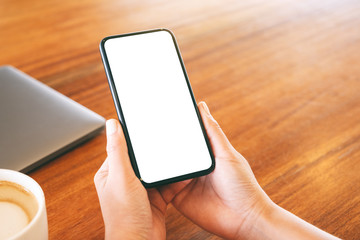 Mockup image of a woman holding black mobile phone with blank desktop screen with laptop and coffee cup on the table