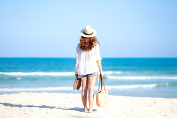 A woman holding bag and shoes while strolling on the beach with the sea and blue sky background