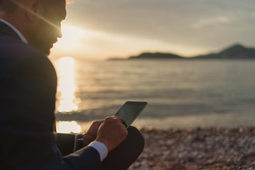 Businessman in a suit using tablet while sitting on the sand of the beach during sunset