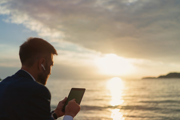 Businessman in a suit using tablet with headphones while sitting on the beach during sunset
