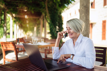 Mature woman using mobile phone while resting at the cafe with laptop computer