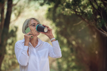 Mature woman talking on mobile phone while resting at the park. Senior lady drink takeaway coffee