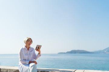 Smiling mature woman having a video chat through tablet on background the sea and blue sky