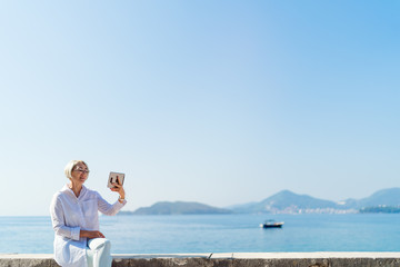 Fototapeta na wymiar Smiling mature woman having a video chat through tablet on background the sea and blue sky