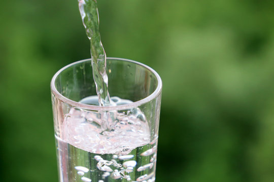 Pouring clean water into drinking glass on green nature background. Concept of health and freshness, thirst, water purification and ecology