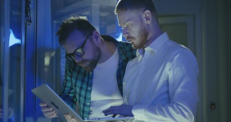 Two male young IT system administrators discuss work using laptop in rack server