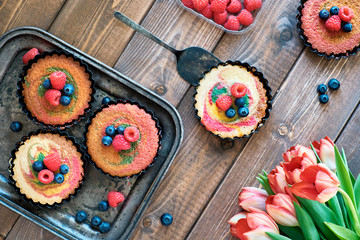 Flat lay with small parrotot cakes decorated with raspberry and blueberry on dark wooden boards with tulips