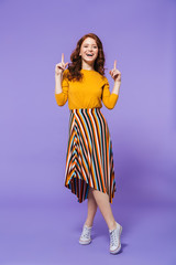 Full length portrait of a pretty young redheaded woman isolated over violet background, pointing finger at copy space