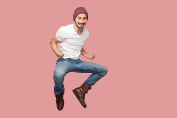 Portrait of happy bearded hipster young man in white shirt and blue jeans with hat jumping, rejoicing and celebrating his victory. indoor studio shot, isolated on pink background copyspace.